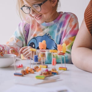 May include: A young girl wearing glasses and a tie-dye shirt builds a tower with colourful candy and toothpicks on a white table.