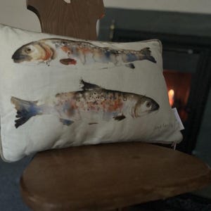 May include: A white throw pillow with a watercolor illustration of two fish. The fish are brown, orange, and white with a light blue background. The pillow is sitting on a wooden chair.