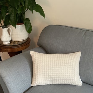 May include: A white cable knit throw pillow on a gray sofa. The pillow is rectangular and has a subtle texture.