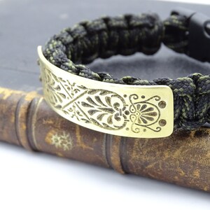 Puede incluir: Pulsera hecha a mano con un cord&oacute;n trenzado negro y verde oliva. Cuenta con una placa met&aacute;lica dorada con un dise&ntilde;o floral grabado. El cierre es negro.
