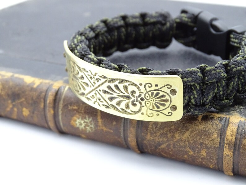 Puede incluir: Pulsera hecha a mano con un cord&oacute;n trenzado negro y verde oliva. Cuenta con una placa met&aacute;lica dorada con un dise&ntilde;o floral grabado. El cierre es negro.