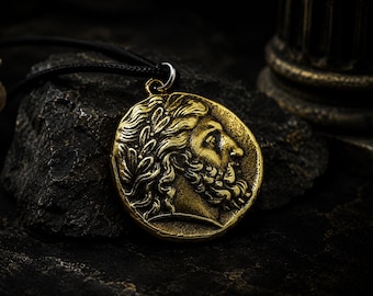 May include: A gold-colored pendant necklace featuring a detailed profile of a man with a beard and laurel wreath. The pendant is circular and attached to a black cord. The background is dark, with a textured rock and a hint of a column.