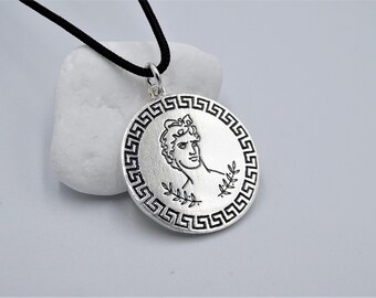 May include: Silver pendant necklace with a black cord. The pendant features a profile of a man's face with a laurel wreath around his head, surrounded by a Greek key border.