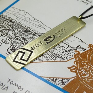 May include: A gold-coloured metal pendant necklace with a rectangular tag featuring a Greek symbol and the text "&Epsilon;&Sigma;&Sigma;&Epsilon;&Tau;&Iota; &Eta;&Mu;&Alpha;&Rho; &Omicron;&Iota;&Eta;&Rho;&Omicron;&Sigma;".