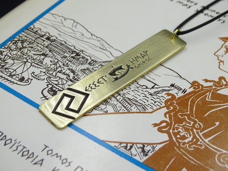 May include: A gold-coloured metal pendant necklace with a rectangular tag featuring a Greek symbol and the text "&Epsilon;&Sigma;&Sigma;&Epsilon;&Tau;&Iota; &Eta;&Mu;&Alpha;&Rho; &Omicron;&Iota;&Eta;&Rho;&Omicron;&Sigma;".
