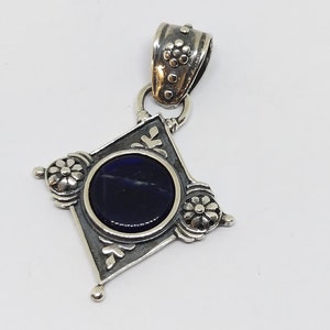 May include: A silver pendant with a dark blue gemstone set in a diamond-shaped frame. The frame is decorated with floral designs and has a silver bail.