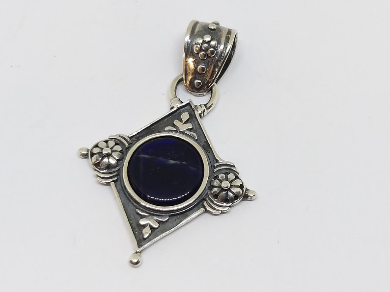 May include: A silver pendant with a dark blue gemstone set in a diamond-shaped frame. The frame is decorated with floral designs and has a silver bail.