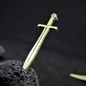 Peut inclure: Un pendentif en forme d'épée de couleur dorée avec un design simple, reposant sur une roche gris foncé.