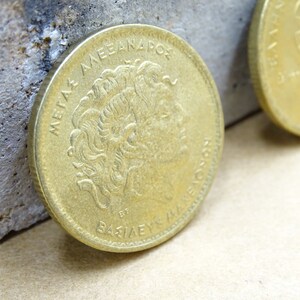 Puede incluir: Una moneda griega de color dorado con el texto "METAXA AEANAPOC ET BAZIAEYC" y un perfil de cabeza humana en un lado.