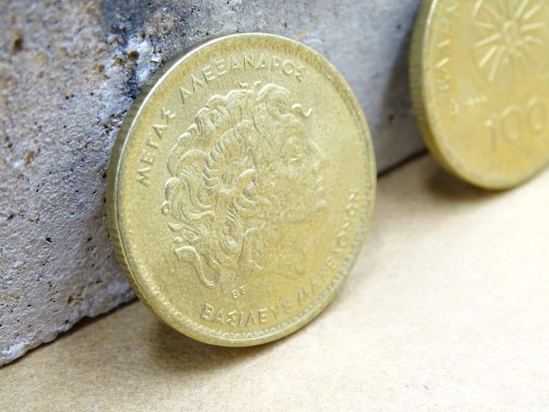 Puede incluir: Una moneda griega de color dorado con el texto "METAXA AEANAPOC ET BAZIAEYC" y un perfil de cabeza humana en un lado.