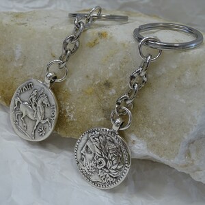 May include: Two silver keychains with round coin-shaped charms. One charm features a depiction of a rider on horseback, the other charm features a profile of a person with a laurel wreath.