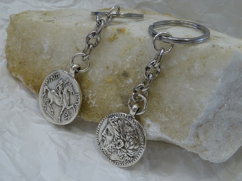 May include: Two silver keychains with round coin-shaped charms. One charm features a depiction of a rider on horseback, the other charm features a profile of a person with a laurel wreath.