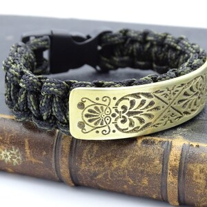 Puede incluir: Pulsera artesanal con un cord&oacute;n tejido negro y verde oliva. La pulsera tiene una placa decorativa de lat&oacute;n con un dise&ntilde;o floral grabado. Un cierre de pl&aacute;stico negro asegura la pulsera.