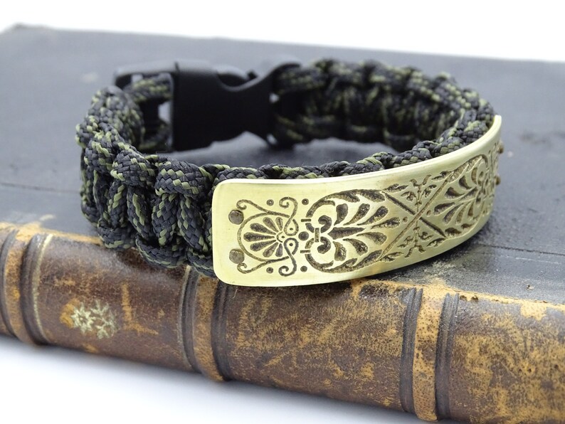 Puede incluir: Pulsera artesanal con un cord&oacute;n tejido negro y verde oliva. La pulsera tiene una placa decorativa de lat&oacute;n con un dise&ntilde;o floral grabado. Un cierre de pl&aacute;stico negro asegura la pulsera.
