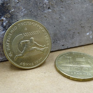 May include: Two gold-colored Greek drachma coins. The first coin features a stylized image of a runner with the text "ATHENS 1997" and "VI. PANATHENAIC GAMES OF ATHLETICS" in Greek. The second coin features a stylized image of the Panathenaic Stadium with the text "100 DRAKHMES".