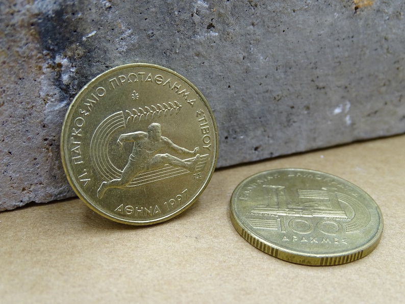 May include: Two gold-colored Greek drachma coins. The first coin features a stylized image of a runner with the text "ATHENS 1997" and "VI. PANATHENAIC GAMES OF ATHLETICS" in Greek. The second coin features a stylized image of the Panathenaic Stadium with the text "100 DRAKHMES".