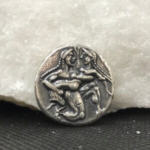 Puede incluir: Una moneda de plata con un relieve detallado de dos figuras abraz&aacute;ndose. Las figuras tienen pelo largo y est&aacute;n representadas en un estilo cl&aacute;sico. La moneda tiene una apariencia desgastada y antigua.