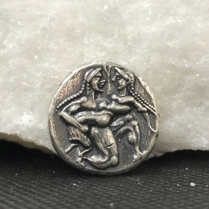 Puede incluir: Una moneda de plata con un relieve detallado de dos figuras abraz&aacute;ndose. Las figuras tienen pelo largo y est&aacute;n representadas en un estilo cl&aacute;sico. La moneda tiene una apariencia desgastada y antigua.