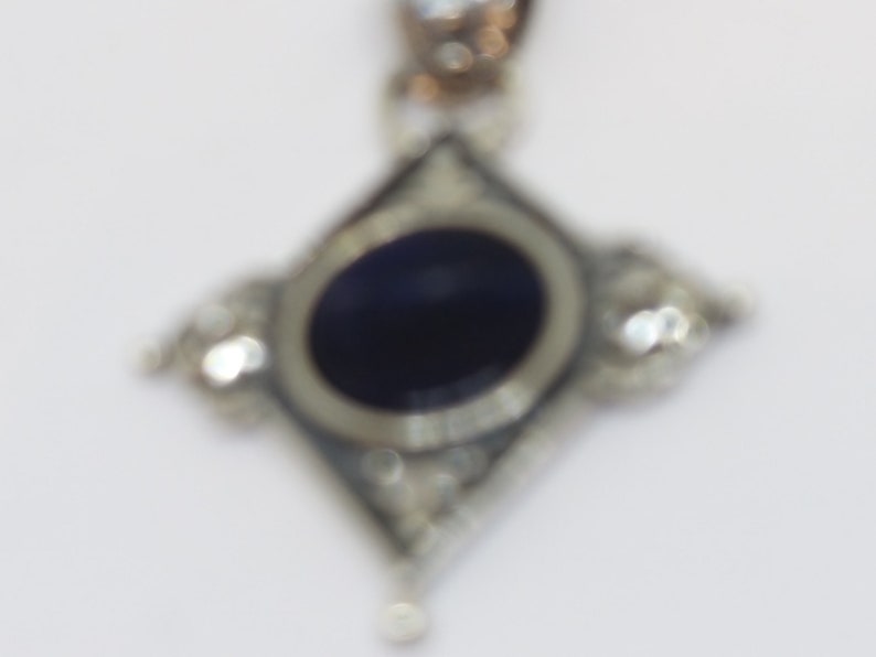 May include: A silver pendant with a dark blue oval gemstone surrounded by smaller clear gemstones. The pendant is in the shape of a diamond.