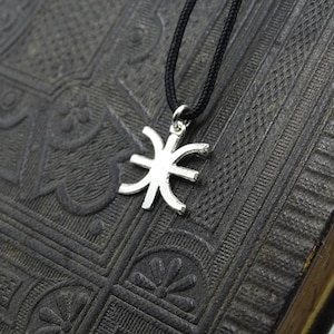 Puede incluir: Un colgante plateado con un diseño único y estilizado cuelga de un cordón negro. El colgante está centrado sobre un fondo oscuro y texturizado, posiblemente un libro o una superficie similar. El diseño del colgante es el punto focal.