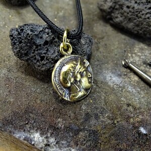 Puede incluir: Un collar con un colgante redondo en forma de moneda de color dorado, que presenta el perfil de dos caras. El collar est&aacute; en un cord&oacute;n negro y descansa sobre una superficie rugosa de color gris oscuro.