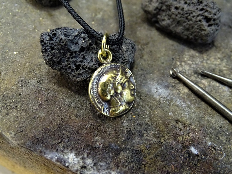 Puede incluir: Un collar con un colgante redondo en forma de moneda de color dorado, que presenta el perfil de dos caras. El collar est&aacute; en un cord&oacute;n negro y descansa sobre una superficie rugosa de color gris oscuro.