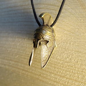 May include: A gold-colored metal pendant in the shape of a Spartan helmet. The pendant is hanging from a black cord.