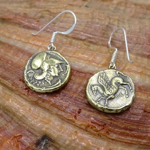 May include: A pair of gold-toned earrings with a coin-shaped design. One earring features a winged horse, while the other features a profile of a person wearing a helmet. The earrings are suspended from silver-toned hooks.
