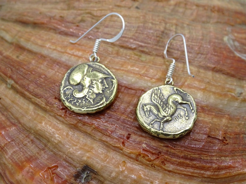 May include: A pair of gold-toned earrings with a coin-shaped design. One earring features a winged horse, while the other features a profile of a person wearing a helmet. The earrings are suspended from silver-toned hooks.
