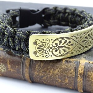 Puede incluir: Pulsera artesanal de paracord con dise&ntilde;o trenzado en negro y verde oliva. Cuenta con una placa decorativa de lat&oacute;n con un patr&oacute;n floral grabado y un cierre de pl&aacute;stico negro.