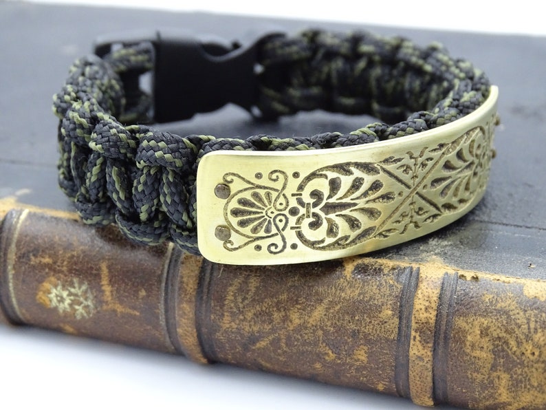 Puede incluir: Pulsera artesanal de paracord con dise&ntilde;o trenzado en negro y verde oliva. Cuenta con una placa decorativa de lat&oacute;n con un patr&oacute;n floral grabado y un cierre de pl&aacute;stico negro.