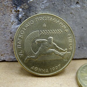 May include: A gold-colored coin with the inscription "VI. PANATHENAIKO PROTASGHMA STIBOY" and "ATHINA 1997" around the edge. The coin features a stylized image of a runner.