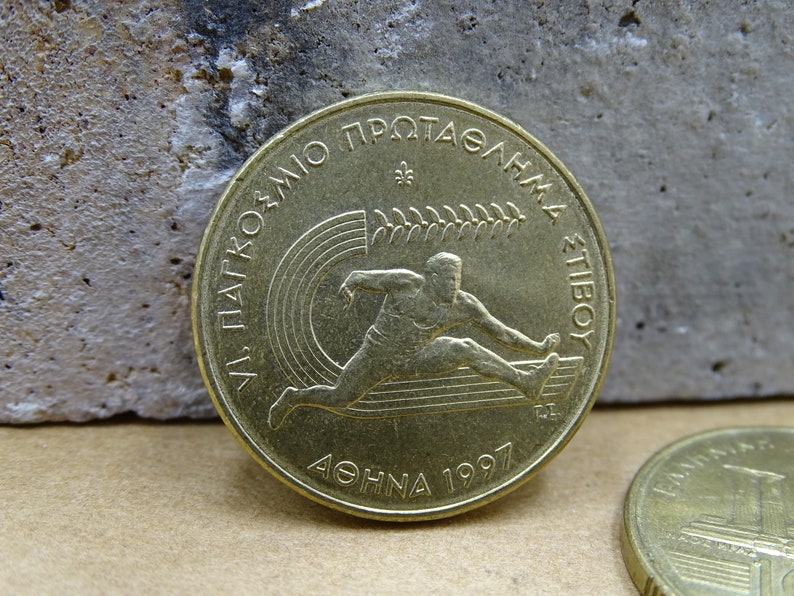 May include: A gold-colored coin with the inscription "VI. PANATHENAIKO PROTASGHMA STIBOY" and "ATHINA 1997" around the edge. The coin features a stylized image of a runner.