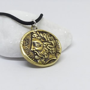 May include: A gold-toned pendant necklace with a round coin-shaped charm featuring a profile of a man with a beard and a laurel wreath. The inscription on the coin reads "PAEION".