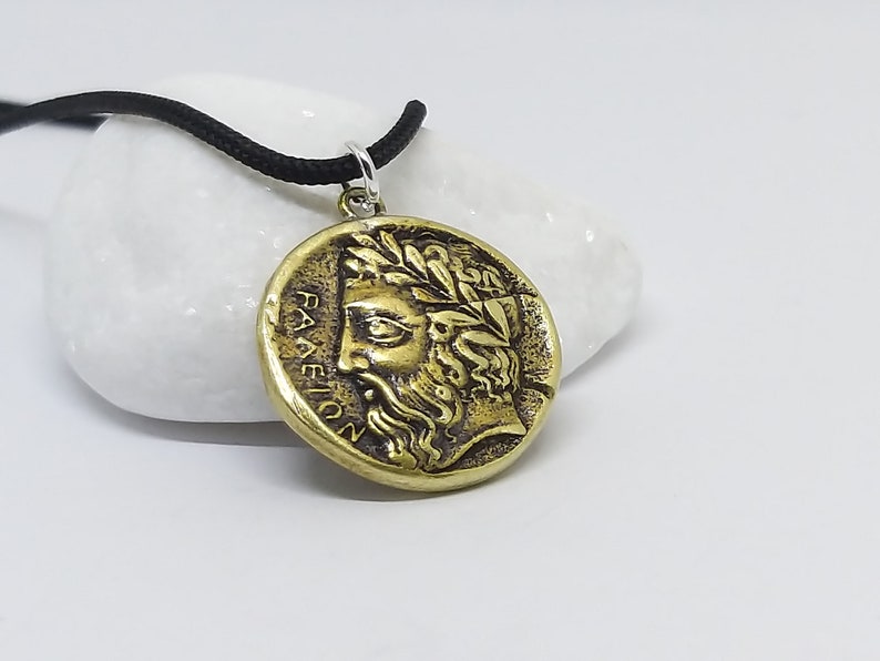 May include: A gold-toned pendant necklace with a round coin-shaped charm featuring a profile of a man with a beard and a laurel wreath. The inscription on the coin reads "PAEION".