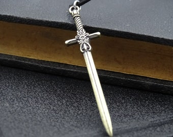Pode incluir: Um pingente de espada de prata com detalhes intrincados no punho e na lâmina. O pingente está pendurado em um cordão preto.