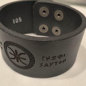 May include: A black leather cuff bracelet featuring four metallic snap buttons, the number 105 printed on the interior, and an engraved circular emblem with a central symbol and the text SAYTON on the exterior surface of the band.