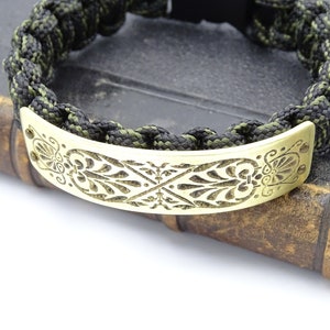 Puede incluir: Una pulsera trenzada con un cord&oacute;n negro y verde oliva. La pulsera presenta una placa met&aacute;lica decorativa, curvada, de color dorado con un dise&ntilde;o floral grabado. La pulsera se muestra sobre un libro antiguo.
