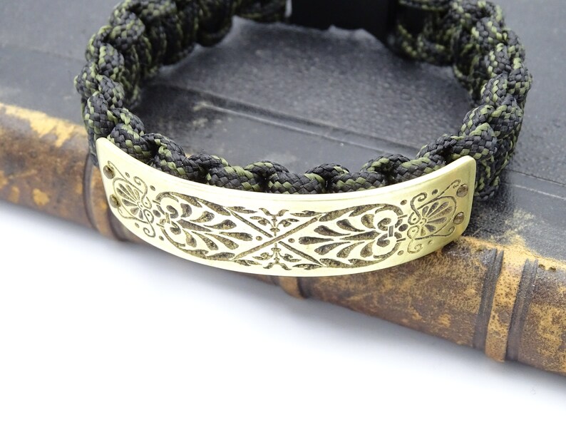 Puede incluir: Una pulsera trenzada con un cord&oacute;n negro y verde oliva. La pulsera presenta una placa met&aacute;lica decorativa, curvada, de color dorado con un dise&ntilde;o floral grabado. La pulsera se muestra sobre un libro antiguo.