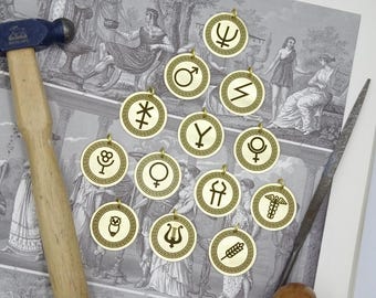 May include: A collection of 14 gold-toned metal charms with various Greek symbols and designs. The charms are circular and feature a Greek key border. The symbols include a lyre, an owl, a wheat stalk, a caduceus, a crescent moon, a star, a sun, a trident, a yin-yang symbol, a female symbol, a male symbol, a double-headed axe, and a letter Y.