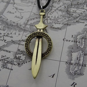 Może przedstawiać: Złoty naszyjnik z zawieszką w kształcie miecza, otoczony okrągłym wzorem. Miecz ma ostry czubek i prosty ostrze. Okrągły wz&oacute;r ma grecki wz&oacute;r klucza.