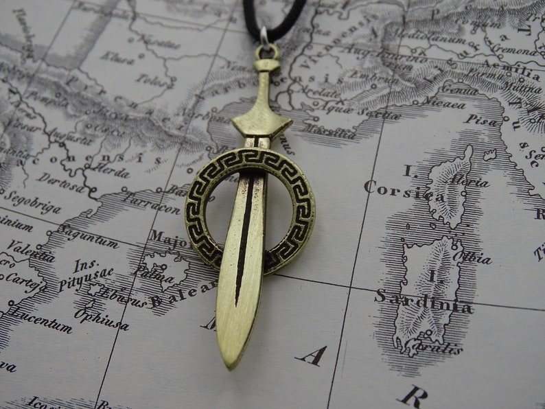 Może przedstawiać: Złoty naszyjnik z zawieszką w kształcie miecza, otoczony okrągłym wzorem. Miecz ma ostry czubek i prosty ostrze. Okrągły wz&oacute;r ma grecki wz&oacute;r klucza.