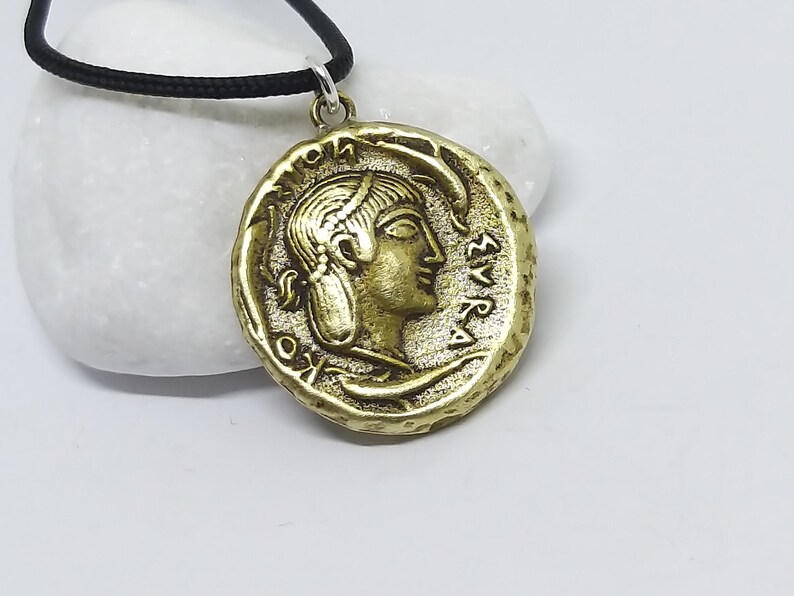 Puede incluir: Un collar con un colgante en forma de moneda griega antigua de color dorado, que representa el perfil de una mujer y letras griegas. El collar est&aacute; en un cord&oacute;n negro.