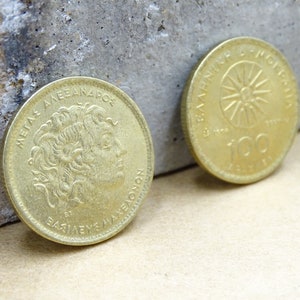 Puede incluir: Dos monedas griegas de dracma de color dorado. La cara anverso presenta un retrato de Alejandro Magno con la inscripci&oacute;n "&Mu;&Epsilon;&Gamma;&Alpha;&Sigma; &Alpha;&Lambda;&Epsilon;&Xi;&Alpha;&Nu;&Delta;&Rho;&Omicron;&Sigma;" y "&Beta;&Alpha;&Sigma;&Iota;&Lambda;&Epsilon;&Upsilon;&Sigma; &Mu;&Alpha;&Kappa;&Epsilon;&Delta;&Omicron;&Nu;&Omega;&Nu;". La cara reverso presenta un sol con rayos y la inscripci&oacute;n "&Epsilon;&Lambda;&Lambda;&Eta;&Nu;&Iota;&Kappa;&Eta; &Delta;&Eta;&Mu;&Omicron;&Kappa;&Rho;&Alpha;&Tau;&Iota;&Alpha;" y "100 &Delta;&Rho;&Alpha;&Chi;&Mu;&Epsilon;&Sigma;".