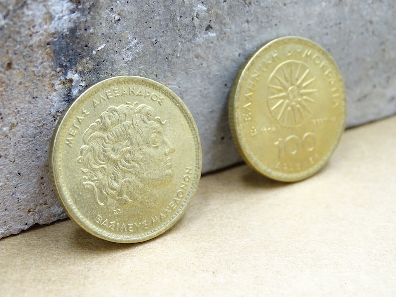 Puede incluir: Dos monedas griegas de dracma de color dorado. La cara anverso presenta un retrato de Alejandro Magno con la inscripci&oacute;n "&Mu;&Epsilon;&Gamma;&Alpha;&Sigma; &Alpha;&Lambda;&Epsilon;&Xi;&Alpha;&Nu;&Delta;&Rho;&Omicron;&Sigma;" y "&Beta;&Alpha;&Sigma;&Iota;&Lambda;&Epsilon;&Upsilon;&Sigma; &Mu;&Alpha;&Kappa;&Epsilon;&Delta;&Omicron;&Nu;&Omega;&Nu;". La cara reverso presenta un sol con rayos y la inscripci&oacute;n "&Epsilon;&Lambda;&Lambda;&Eta;&Nu;&Iota;&Kappa;&Eta; &Delta;&Eta;&Mu;&Omicron;&Kappa;&Rho;&Alpha;&Tau;&Iota;&Alpha;" y "100 &Delta;&Rho;&Alpha;&Chi;&Mu;&Epsilon;&Sigma;".