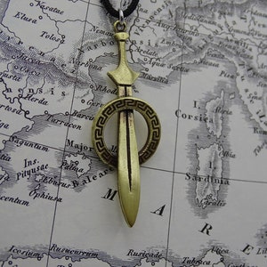 Może przedstawiać: Złoty naszyjnik z zawieszką w kształcie miecza z okrągłym wzorem greckiego klucza. Zawieszka znajduje się na tle mapy.