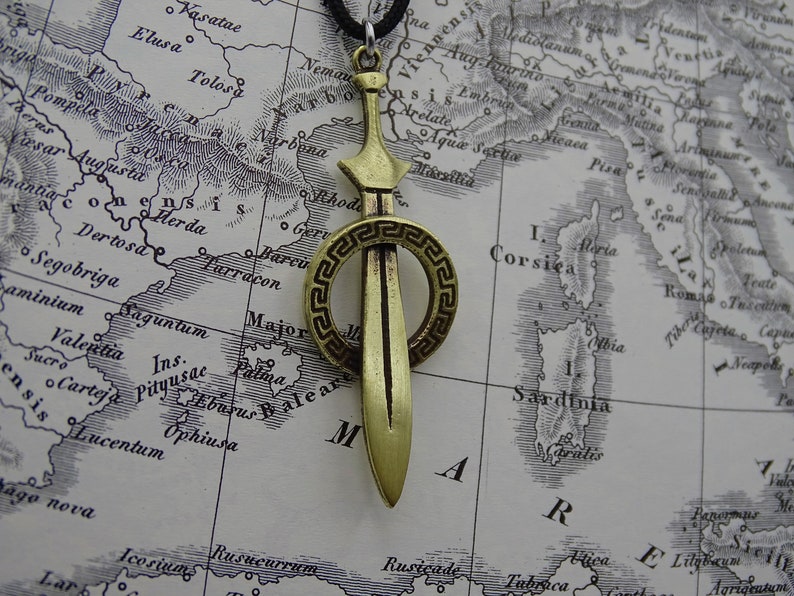 Może przedstawiać: Złoty naszyjnik z zawieszką w kształcie miecza z okrągłym wzorem greckiego klucza. Zawieszka znajduje się na tle mapy.