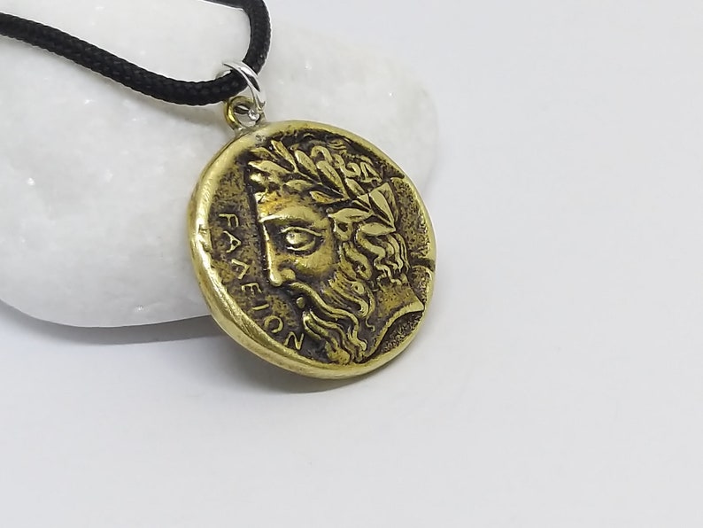 May include: A gold-toned pendant necklace with a round coin-shaped charm featuring a detailed profile of a man with a laurel wreath. The charm has the inscription "FALEION" around the edge.