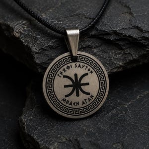 May include: A circular silver-toned pendant on a black cord necklace. The pendant features a central black symbol, Greek-style lettering reading "ΓΝΩΘΙ ΣΑΥΤΟΝ" and "ΜΗΔΕΝ ΑΓΑΝ", and a decorative Greek key border pattern around the outer edge of the metal disc.