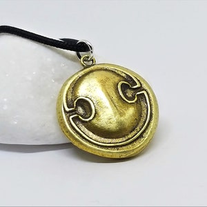 May include: A gold-colored pendant necklace featuring a round shield with a stylized design. The shield has a raised, textured surface and a black cord.